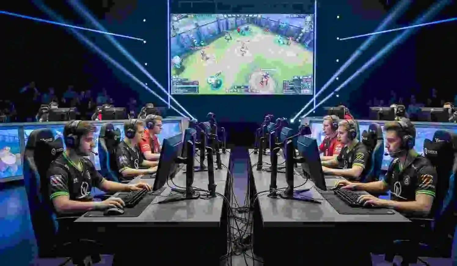 Two teams of professional esports players competing in a live gaming tournament, seated at computer setups with headsets, under vibrant stage lighting and a large gameplay screen in the background – 2025 esports event.